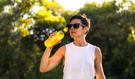Young beautiful woman drink water from the plastic bottle in a park after training, short black hair, sunglasses and shorts, sport outdoors in summer, slim fitness girl in sportswear, female on workoutの写真素材