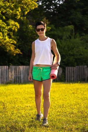 Young beautiful woman with yoga mat in a park, short black hair, sunglasses and shorts, sport outdoors in summer, slim fitness girl in sportswear, female on workout trainingの写真素材