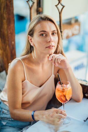 Beautiful young woman in cafe with cold cocktail in hot summer day. Long blonde hair, round earrings and light beige top.の写真素材