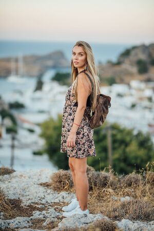 Attractive girl with long blonde hair in flower dress with leather backpack in white sneakersの写真素材