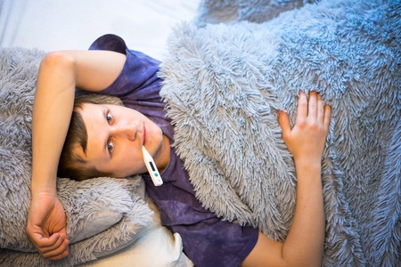 A young man lies under a blanket in bed with discomfort. Check the temperature of the thermometer.の写真素材