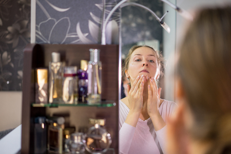 Young woman applies cream on face in front of mirrorの写真素材