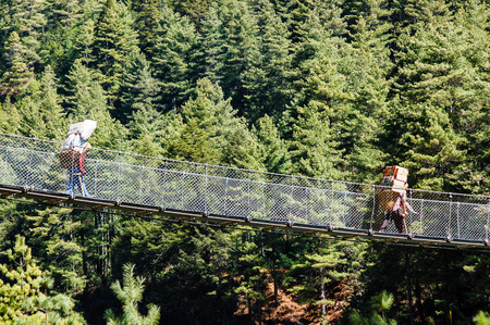 Everest Region, Nepal - March 12, 2010: Two porters carrying heavy stuffs on backs are crossing a canyon by a rope bridge during the everest trekking route.のeditorial素材