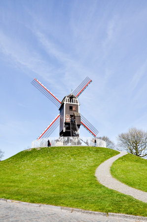 Bruges, Belgium - April 14, 2010: An old traditional wooden windmill on the hill with cobblestone walkway among green grass on a bright blue sky in Bruges.のeditorial素材