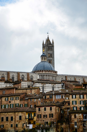 View of Siena's Duomo and campanile, Italian gothic architecture decorated in black and white marble, with medieval town on hillside in foreground and white sky.のeditorial素材