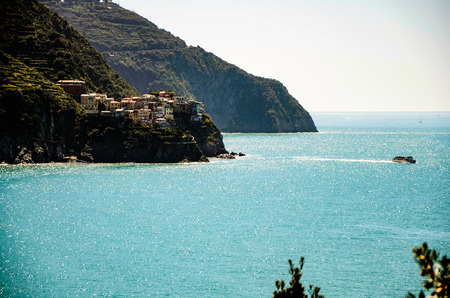 View of traditional colorful Italian houses of Manarola on a cliff at Ligurian sea from Corniglia, Cinque Terre, Italy, with a mountain backdrop and a passenger ferry sailing on the sparkling blue sea on bright sunshine.の写真素材
