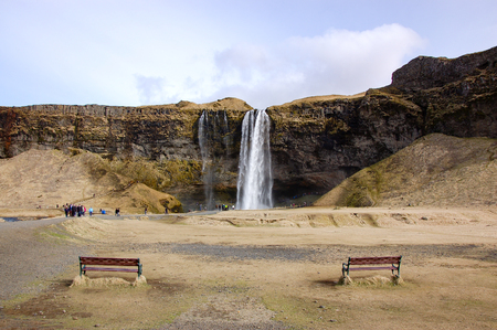 Seljalandsfoss, the waterfall in south Iceland, water falls from the rocky cliff with bright sky and two benches on dry grass in foreground in early spring in horizontal view.の写真素材