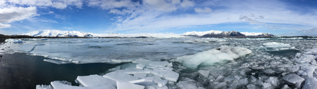 Panorama of cracking ice floe floating on turquoise lake, Jokulsarlon Glacier Lagoon, natural phenomenon with snow-capped mountain backdrop on bright blue sky in southeast Iceland.の写真素材