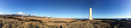 Panorama view of the white lighthouse at Snaefellsnes peninsula, west coast of Iceland with a backdrop of snow-capped mountain and grassland, black lava rock surronded, on blue sky sunshine.の写真素材