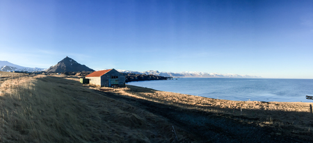 Panorama view of a wooden farmhouse located on the dry pastures at the blue seaside with a backdrop of mountains in a small village, Arnarstapi, Snaefellsnes peninsula, west coast of Iceland  on blue sky sunshine.の写真素材