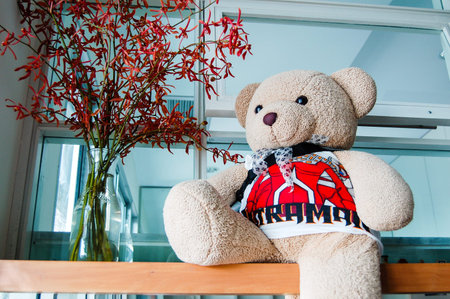 Teddy bear wearing a tank top, sitting on a wooden table beside the red orchid in a glass vase in...