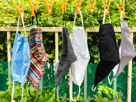 Different style cloth masks lined up for dry in the sun which gripped by orange plastic clothespins with green plastic mesh, steel fence and tree in background.の写真素材