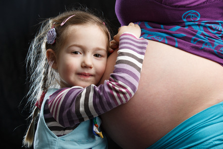 Adorable little girl embracing the belly of a highly pregnant mother, in expectation of a siblingの写真素材