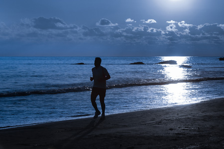 Silhouette of a fit runner in moonlight on a sandy beach in the night. Active lifestyle, vacation recreation, outdoor activity concept.の写真素材