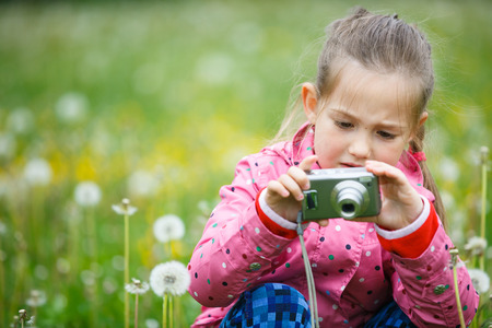 Little curious girl exploring nature with her camera, photographing up-close and kneeling in a dandelion meadow. Active lifestyle, curiosity, pursuing a hobby concept.の写真素材