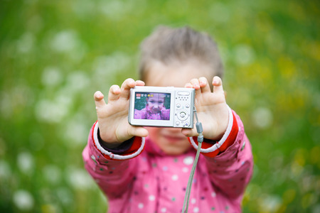 Little cheerful girl making a selfie with digital camera, enjoying her time on a dandelion meadow. Active lifestyle, curiosity, pursuing a hobby, technology and kids  concept.の写真素材