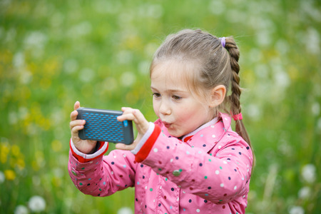 Little curious girl photographing with her smart phone, exploring nature and standing in a dandelion meadow. Active lifestyle, curiosity, pursuing a hobby concept.の写真素材
