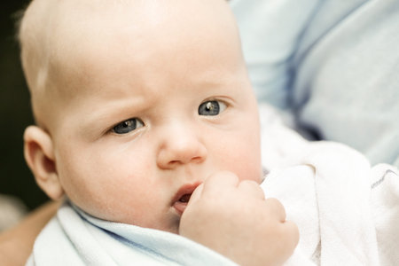 Unique blue-eyed baby boy looking at camera, safely resting in mothers arms in a baby sling wrap,...