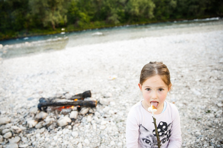 Little girl eating roasted marshmallow by a self-made campfire on camping trip by the mountain river. Active natural lifestyle, childhood concept.の写真素材
