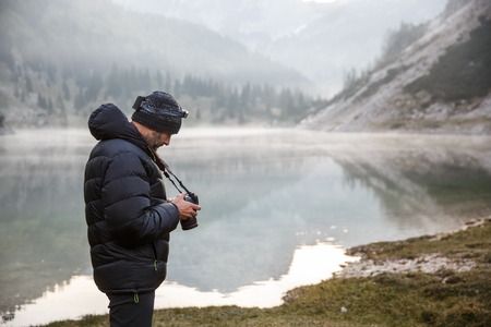 Photographer on assignment, holding a camera, checking photos after photographing in the morning by a mountain lake with winter mist covered surface.の写真素材