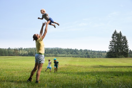 Mother playing with children, throwing a toddler in air, laughing and playing, and her older son and daughter jumping and running on a meadow. Active lifestyle, family time, modern parenting concept. Mother playing with children, throwing a toddler in air, laughing and playing, and her older son and daughter jumping and running on a meadow. Active lifestyle, family time, modern parenting concept.