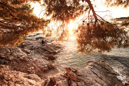 Sunlit rocky beach at sunset with mother and son sitting on shore, cuddling, having fun and enjoying their bonding time together. Family values, travel, tranquility and serenity concept.の写真素材
