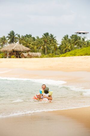 Mother enjoying a tropical beach, playing in the waves with her children. Travelling with children, beach hopping, family time, beach fun concept. の写真素材