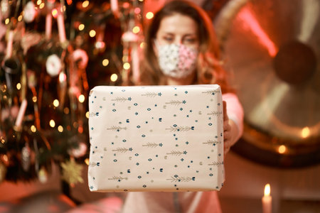 Woman with facial mask holding, giving a wrapped Christmas present. Present delivery, social distancing, Christmas spirit concept.の写真素材