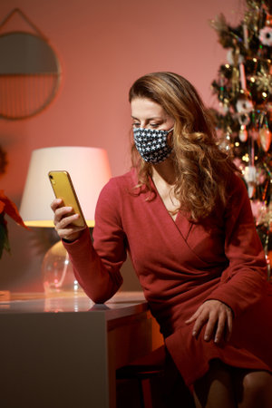 Elegant woman in a red cocktail dress, wearing protective face mask by the christmas tree, alone, lonely, talking and writing on the cell phone. Holiday in quarantine concept.の写真素材