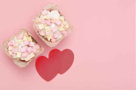 two freshly baked cupcakes covered with sweet icing and heart-shaped marshmallows and two red hearts on a pink background. place for text. Valentine's dayの写真素材