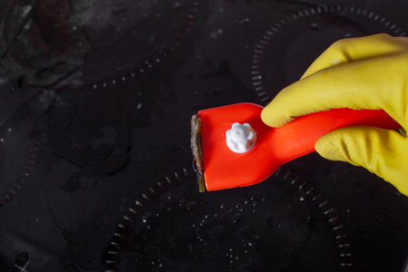 Scraper for cleaning glass ceramics in the hand with rubber glove against the background of a plate...