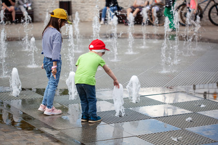 girl and Little boy plays in the square between the water jets in the city fountain at sunny summer day. Active summer leisure for kids in a big city.の写真素材
