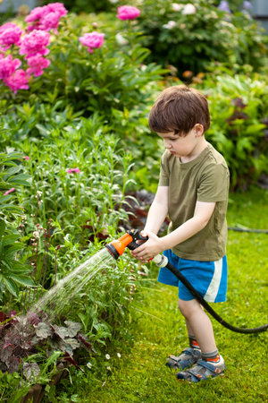 Cute adorable caucasian brown-haired toddler boy enjoy having fun watering garden flower and lawn with hosepipe sprinkler at home backyard on sunny day. Child little helper learn gardening at summer outdoorの写真素材