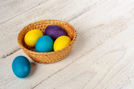 Colored easter eggs in straw basket on wooden white shabby backgroundの写真素材