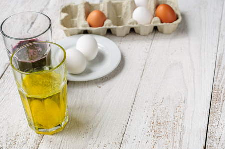 On wooden white shabby background are eggs in carton, eggs on plate and two glasses of food dye with eggsの写真素材