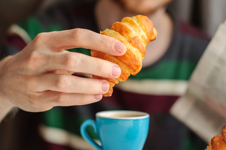 Blurred young man eats his breakfast, coffee, croissant and reads newspaperの写真素材