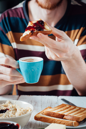 Young man with beard eating coffee and toasts with butter and jam for breakfastの写真素材
