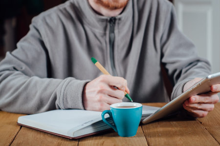 Young man with beard writing notes using tablet pc with cup of coffee