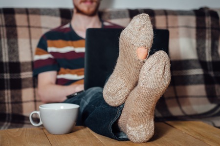 Young man with beard working with laptop in holey socks on tableの写真素材