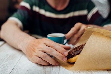Man hands eating chocolate doughnut with coffee on wooden tableの写真素材
