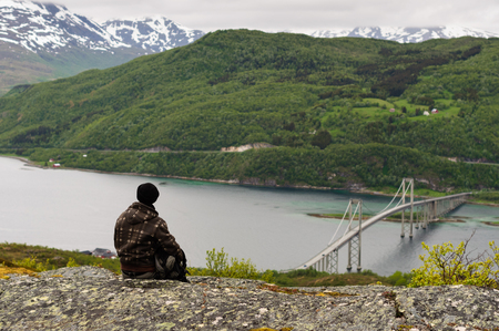 Young man backpacker sitting on mountain peak and looking at fjord in Lofoten, Norwayの写真素材