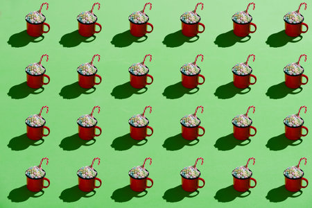 Red enamel mug full of marshmallows and candy cane on green background. Hard light. Pattern