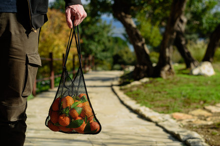 Man holding reusable mesh shopping bag with fresh just picked oranges. Zero waste concept. Sunny day