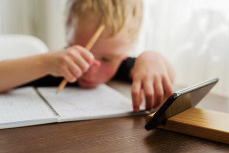 Little sad and tired boy learning at home. Watching webinar by mobile, writing in notebook