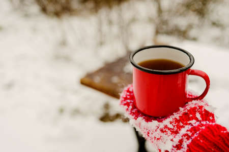 Red mug with hot coffee or tea drink on snow in winter. Red knitted mittens on handsの写真素材