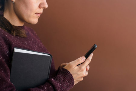 Woman in brown sweater using smartphone. Holding book