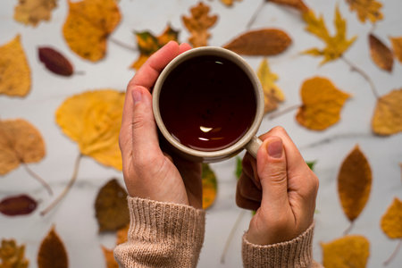 Cup of black tea with autumn leaves on marble background. Woman hands in beige sweater