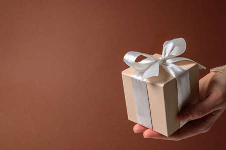 Beige gift box with white bow in woman hands on brown backgroundの写真素材