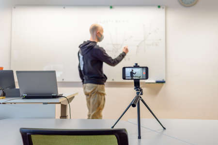 Teacher in face mask in classroom giving online lesson by phone videoの写真素材