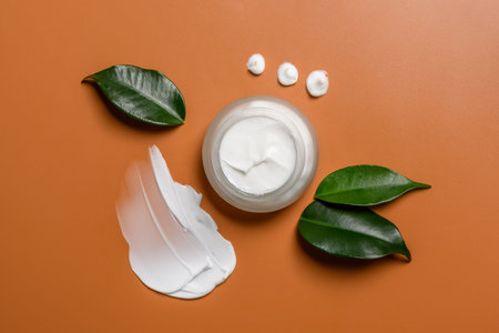 Glass skin cream jar on brown background with smear or swatch and leaves